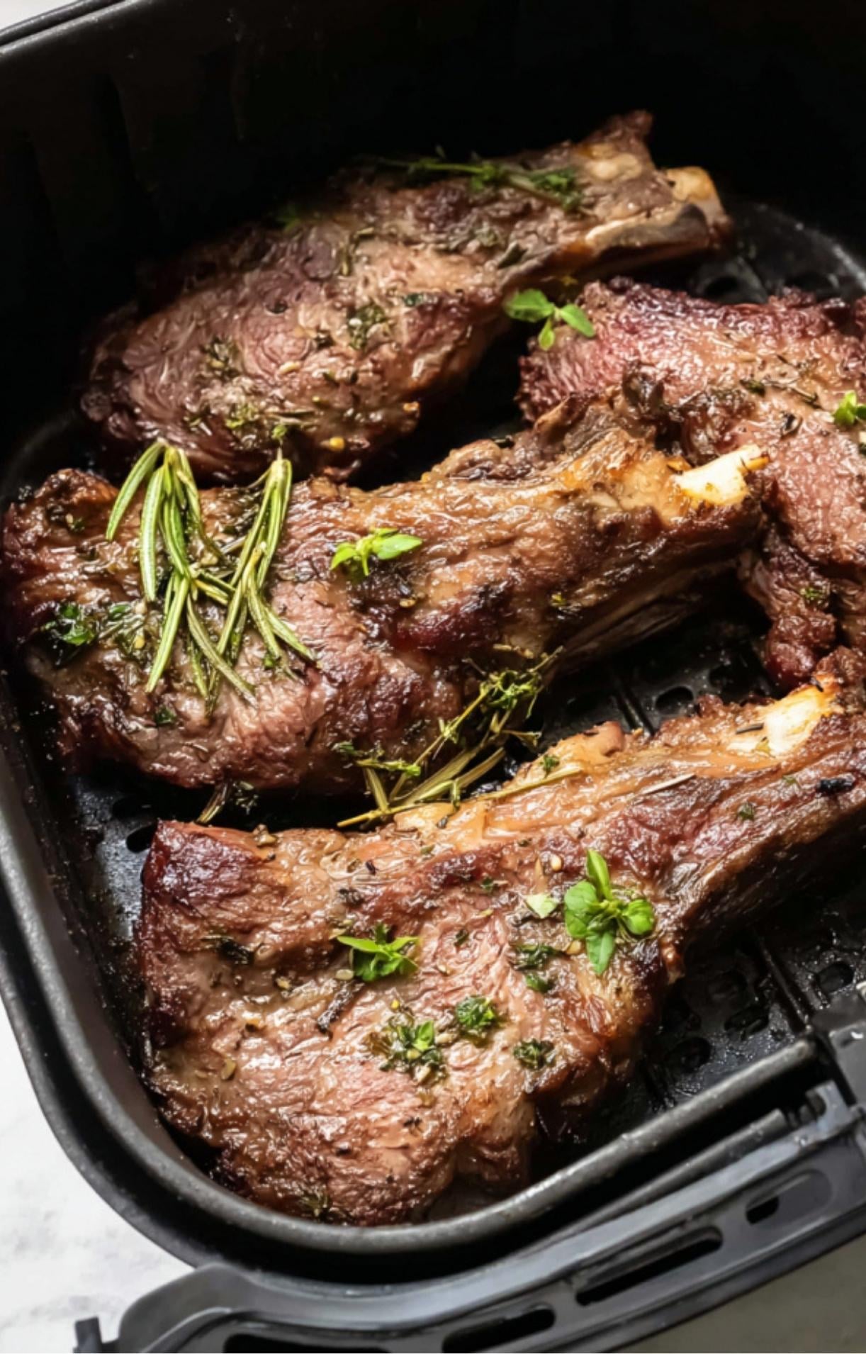 Lamb shoulder chops halfway through cooking in the air fryer, topped with fresh sprigs of rosemary and thyme.