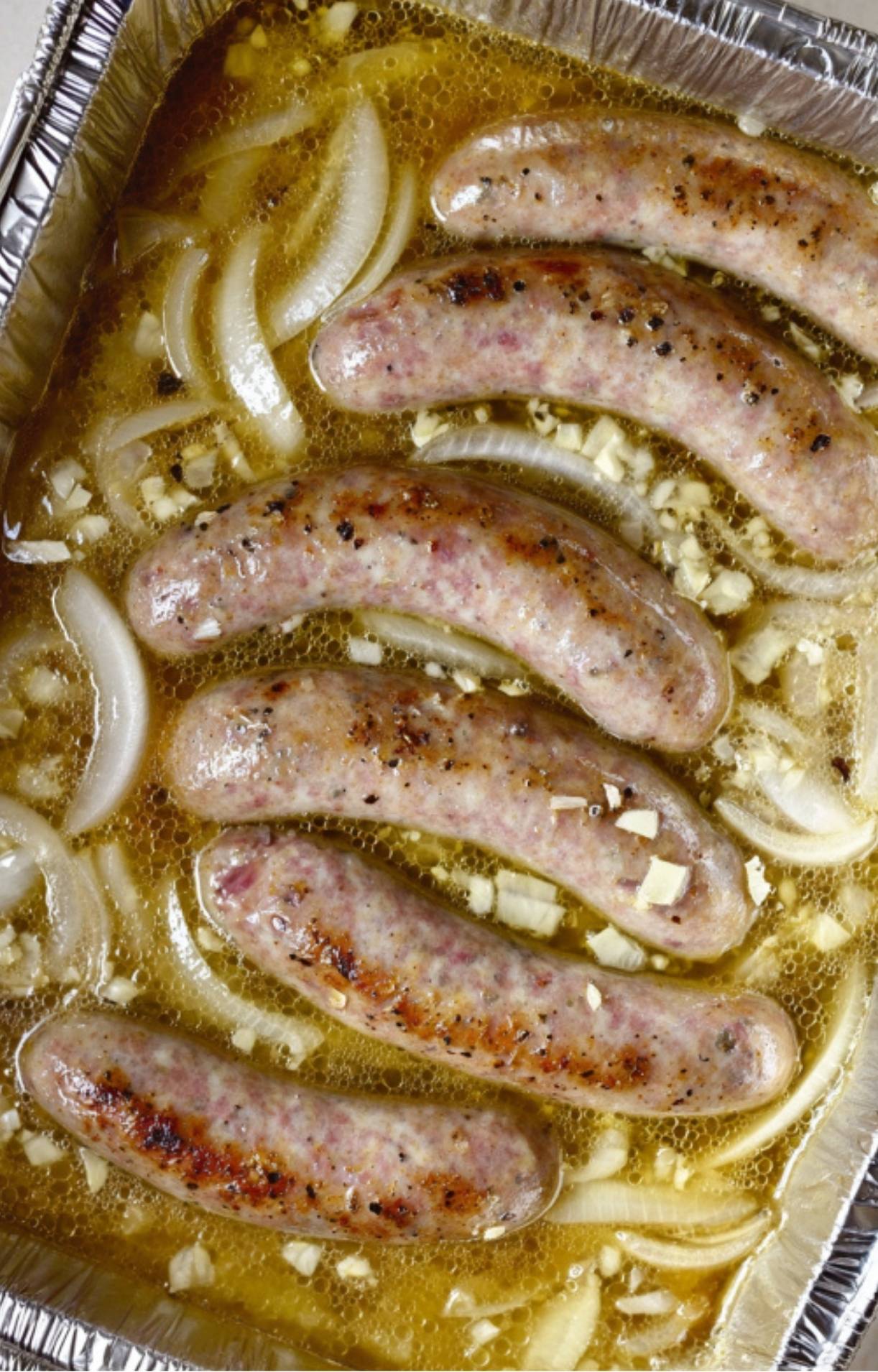 Bratwurst sausages braising in beer with sliced onions and minced garlic in a silver foil tray.
