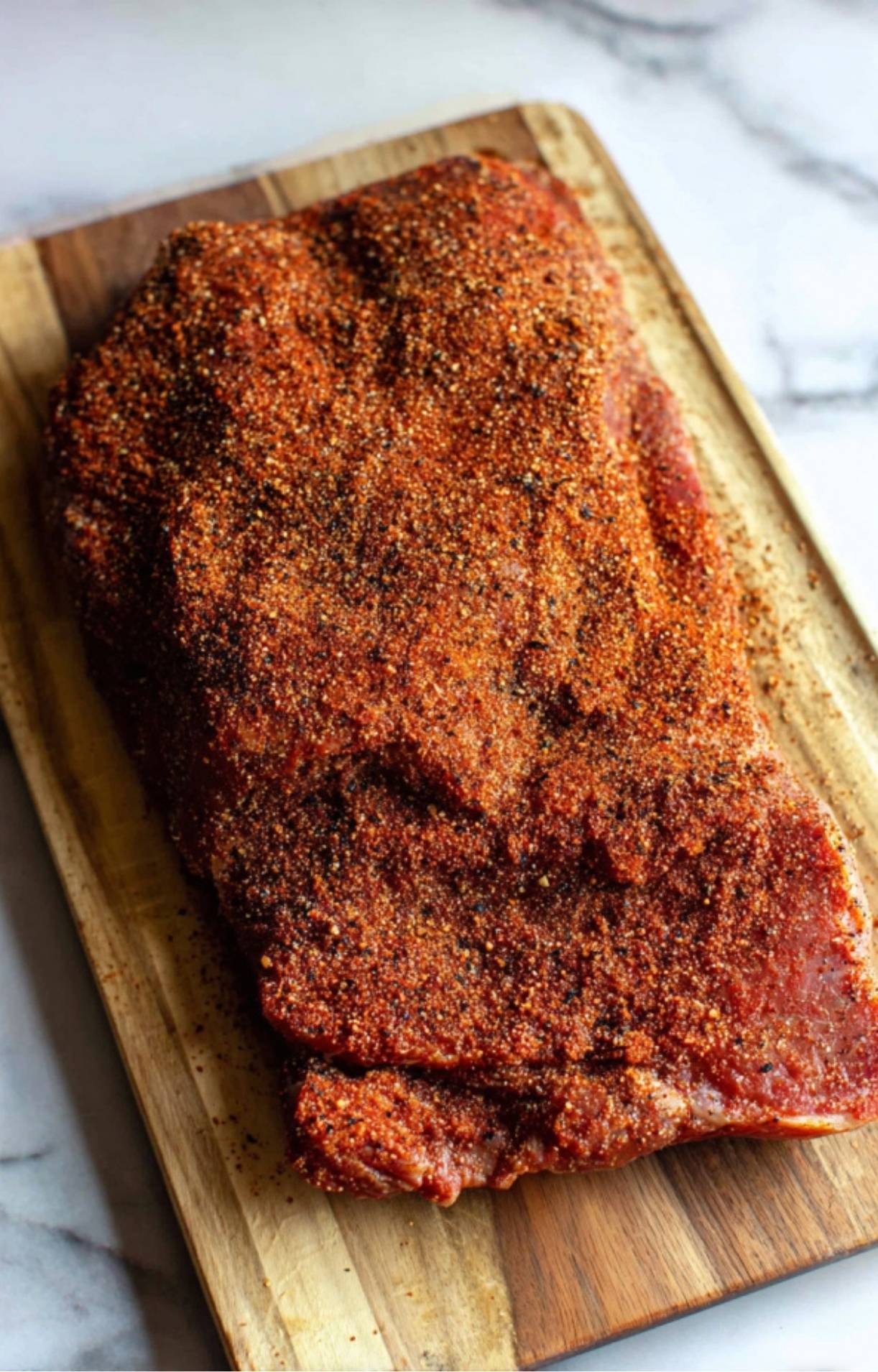 A raw beef brisket resting on a wooden board, completely coated in a reddish-brown spice rub for Electric Smoker Beef Brisket.