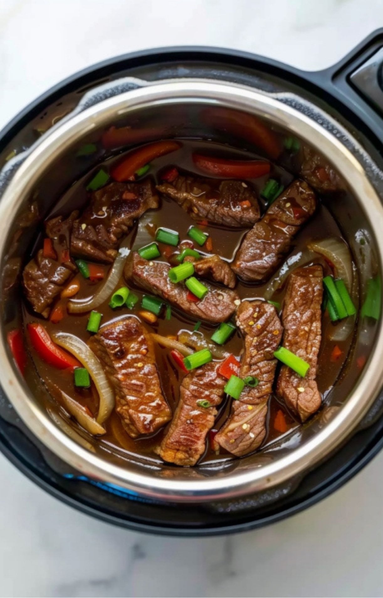A stainless steel Instant Pot bowl containing cooked beef strips, red bell peppers, and onions in a rich brown glaze, freshly topped with sliced green onions for a vibrant garnish.