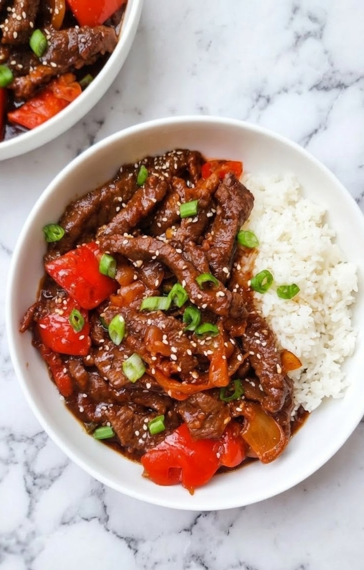 A top-down view of a white bowl filled with Beijing beef stir-fry featuring tender steak strips, red bell peppers, and onions in a dark savory sauce, served alongside white rice and garnished with green onions and sesame seeds.