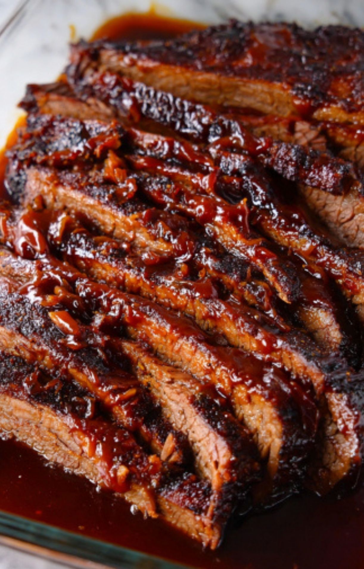 A close-up view of tender, juicy slices of cooked beef brisket resting in its own dark juices in a glass baking dish.