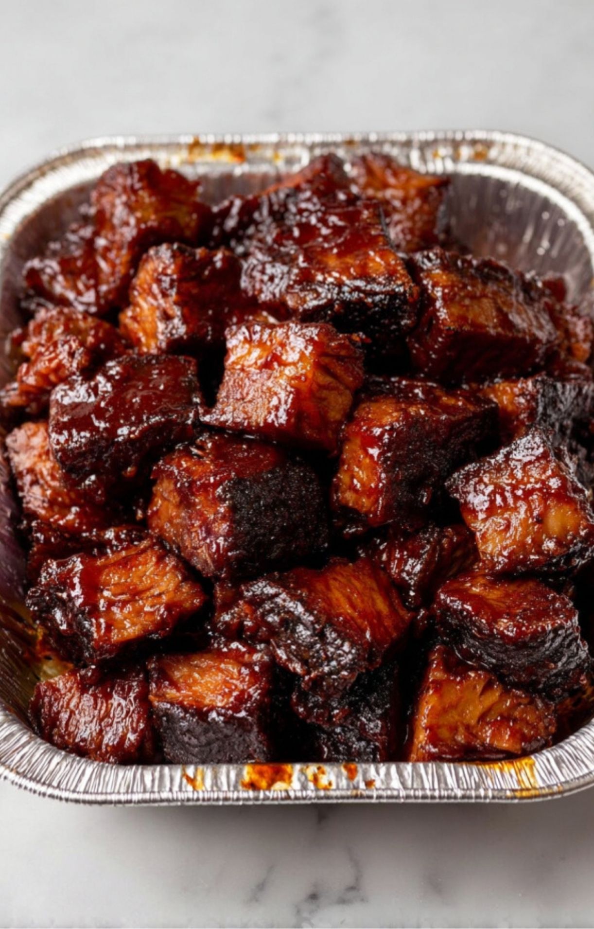 A top-down view of sticky, caramelized Smoked Beef Brisket Burnt Ends piled high in a disposable aluminum pan.