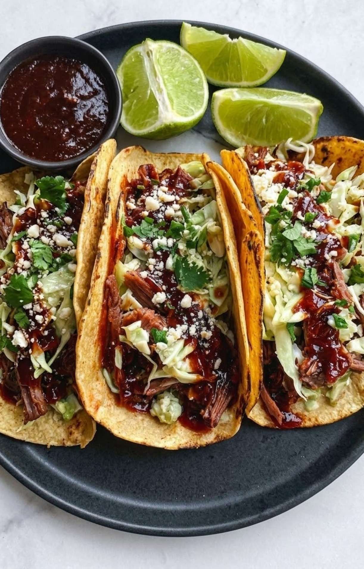 Three tacos filled with shredded brisket, slaw, and cotija cheese served with lime wedges and salsa.