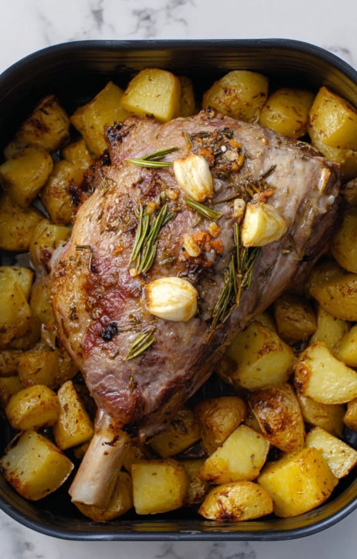 A partially cooked leg of lamb topped with garlic cloves and rosemary, surrounded by golden-brown potatoes in an air fryer basket.