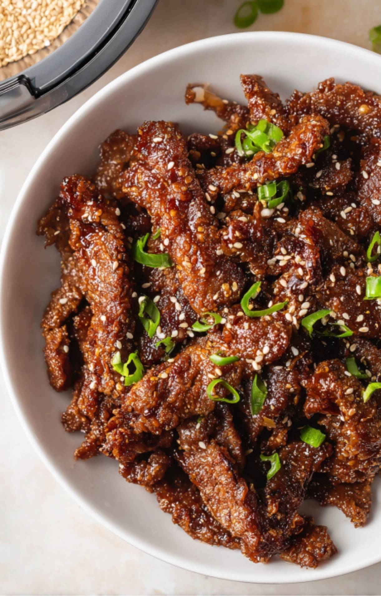 A top-down view of a white bowl filled with crispy, glazed Air Fryer Beijing Beef strips topped with toasted sesame seeds and sliced green onions.