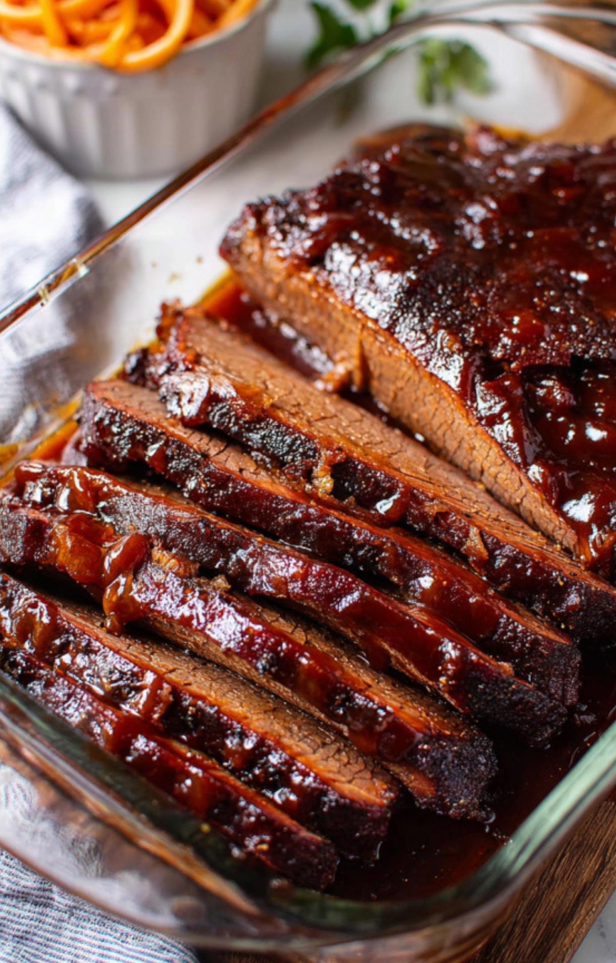 A close-up, angled shot of a glass baking dish filled with thick, succulent slices of oven-smoked beef brisket, dripping in a rich, dark barbecue glaze with a side of curly fries visible in the blurred background.