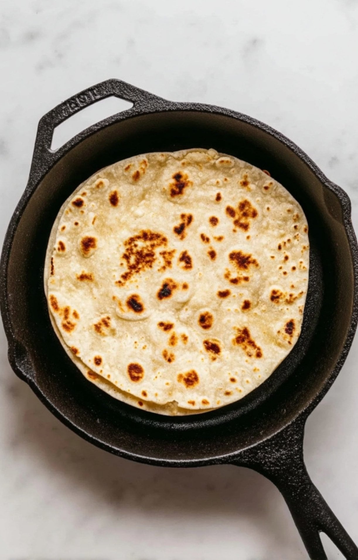 A flour tortilla being toasted and charred in a black cast-iron skillet over high heat.