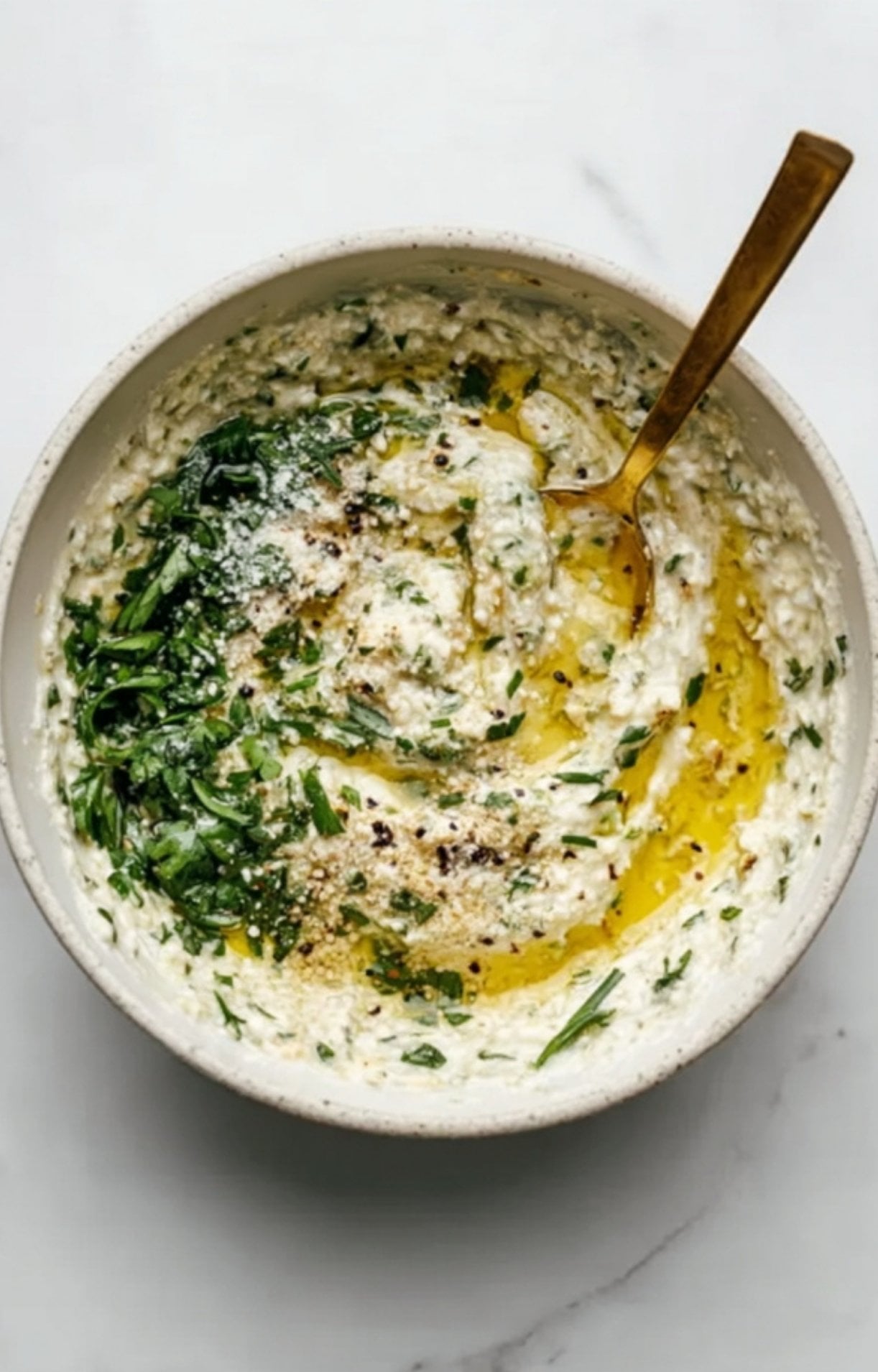 A white bowl containing a coarse mixture of chopped green herbs, olive oil, and seasonings being stirred with a gold spoon.