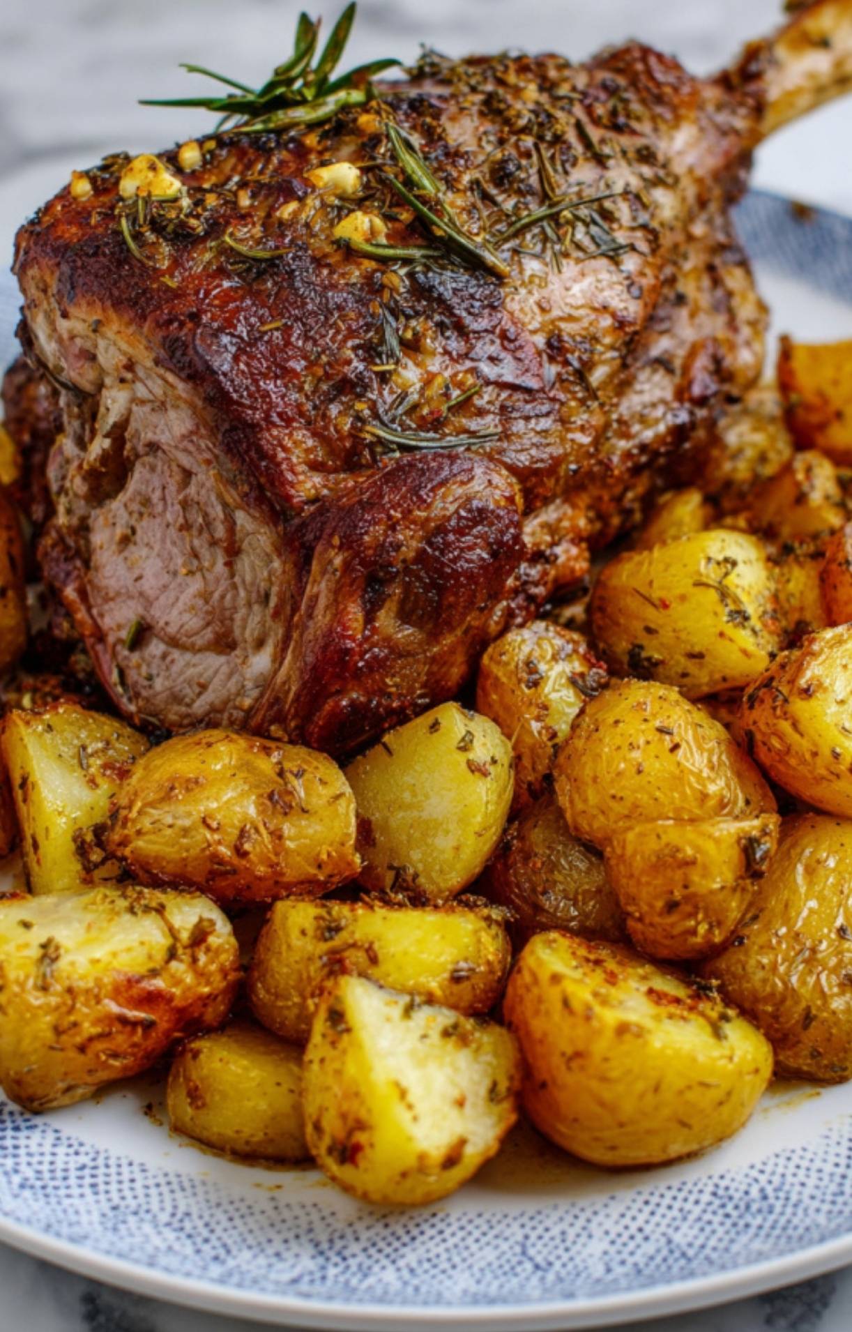 A whole roasted leg of lamb with rosemary garnishes served on a blue and white patterned plate with a side of crispy potatoes.