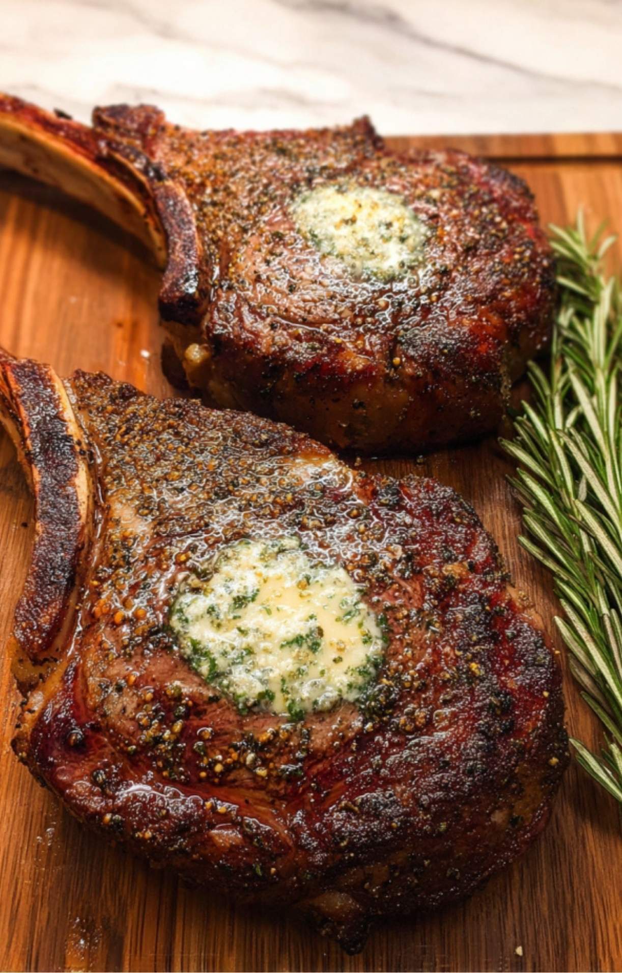Two whole smoked tomahawk steaks resting on a wooden cutting board, topped with melting herb compound butter and fresh rosemary.