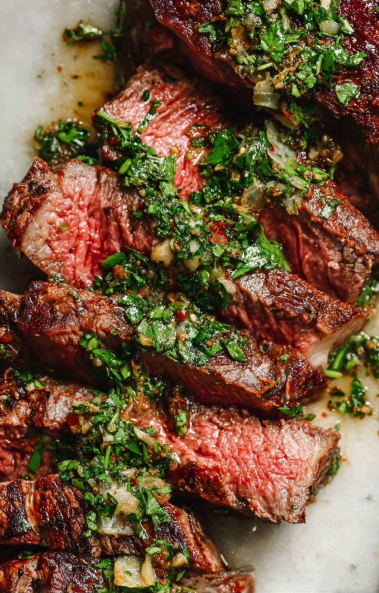 A detailed close-up of juicy, pink medium-rare steak slices glistening with oil and topped with a vibrant green garlic and herb sauce.