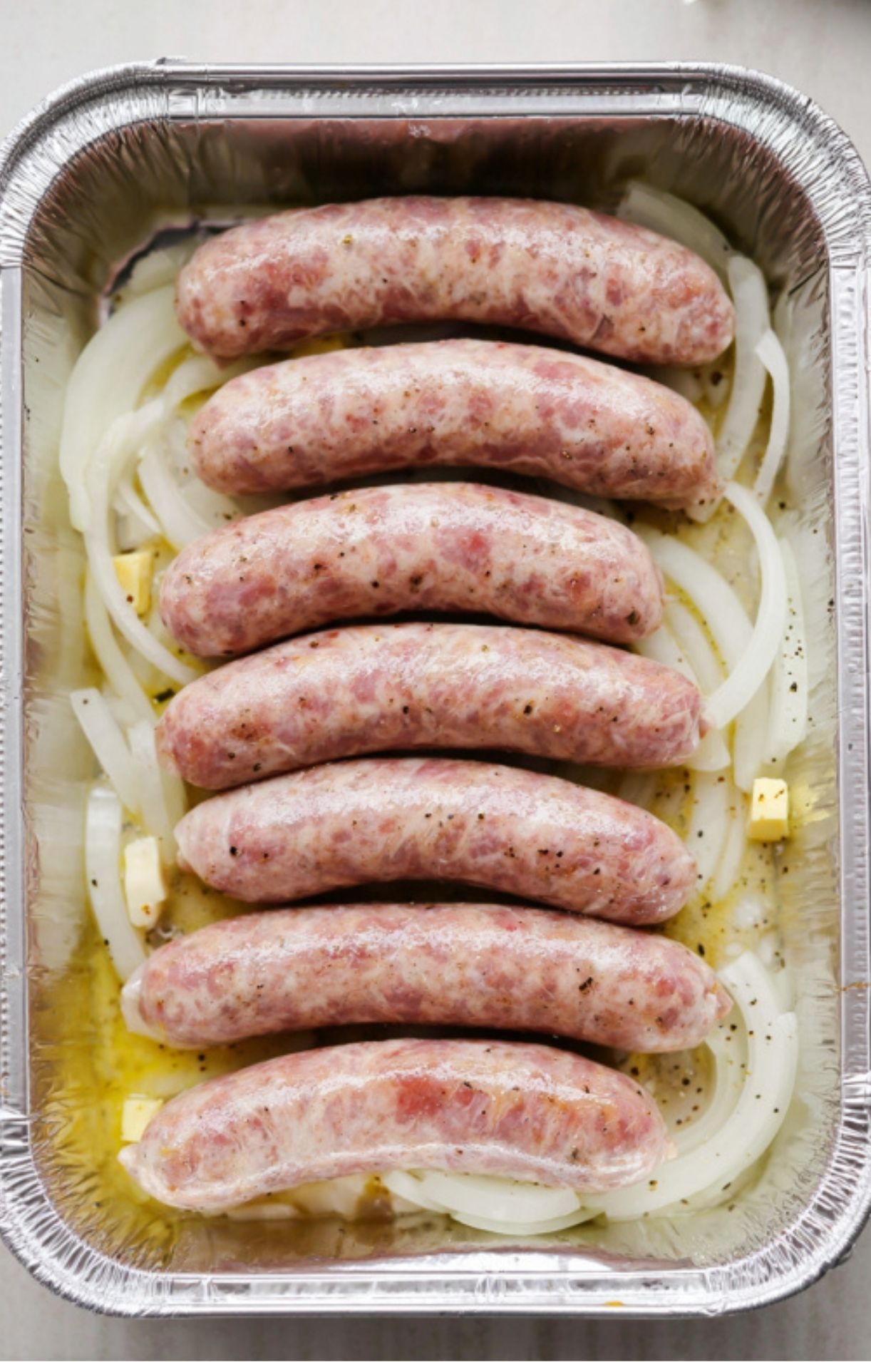 Six raw bratwurst links nestled on top of sliced onions and butter in a disposable foil roasting pan.
