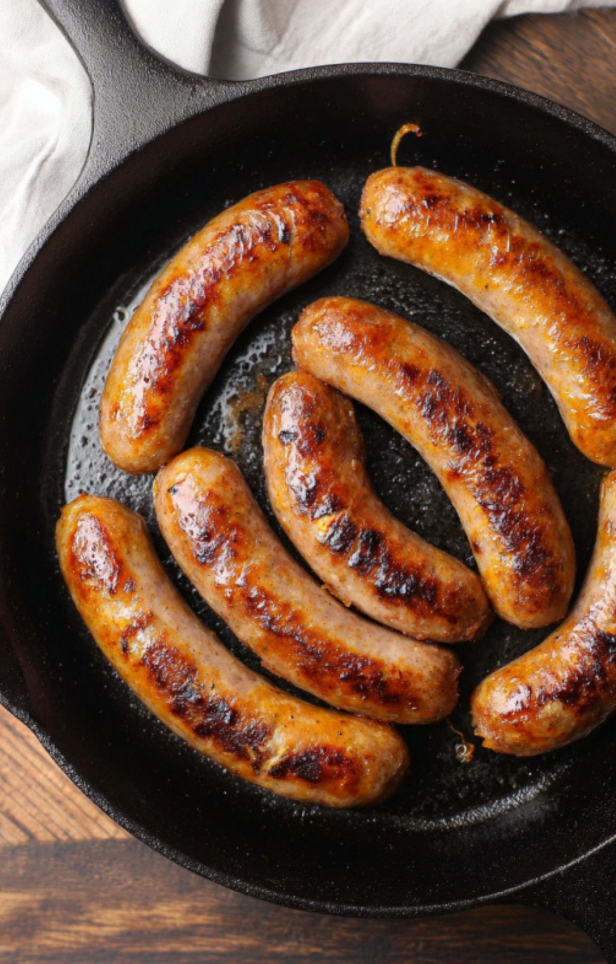 Six bratwurst sausages browning in a hot cast-iron skillet, showing a deep golden-brown sear on the outside.