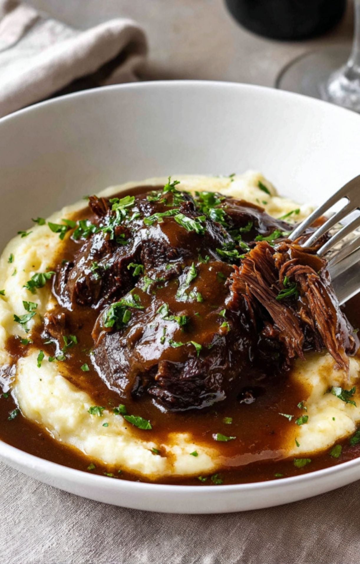 A fork pulling apart a piece of extremely tender slow cooker beef cheek served over a bed of creamy garlic mashed potatoes and topped with a thick, dark brown gravy and chopped parsley.