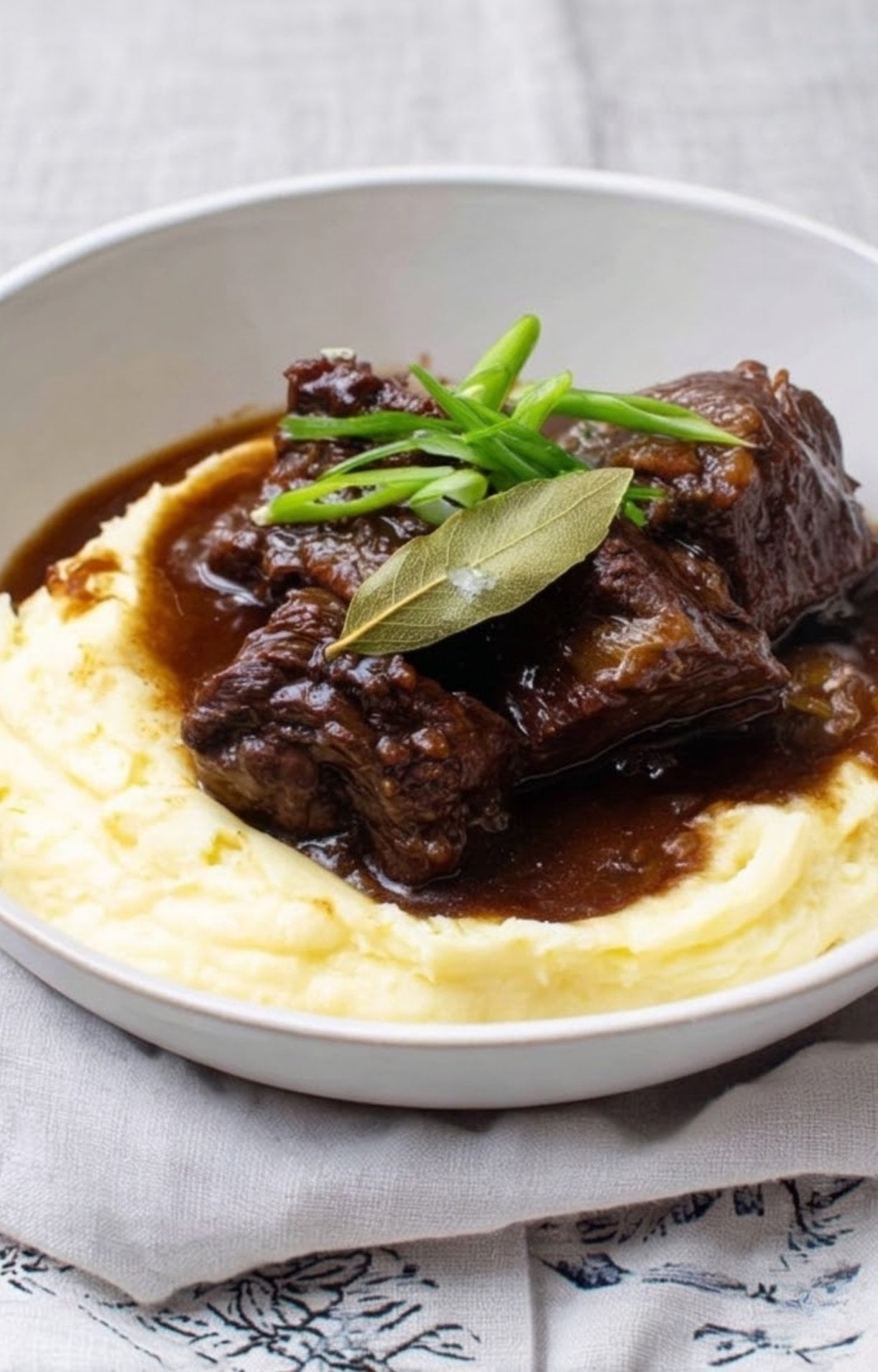 A comforting bowl of slow cooker beef cheeks served over smooth mashed potatoes, garnished with green onions and a single bay leaf in a rich, glossy gravy.