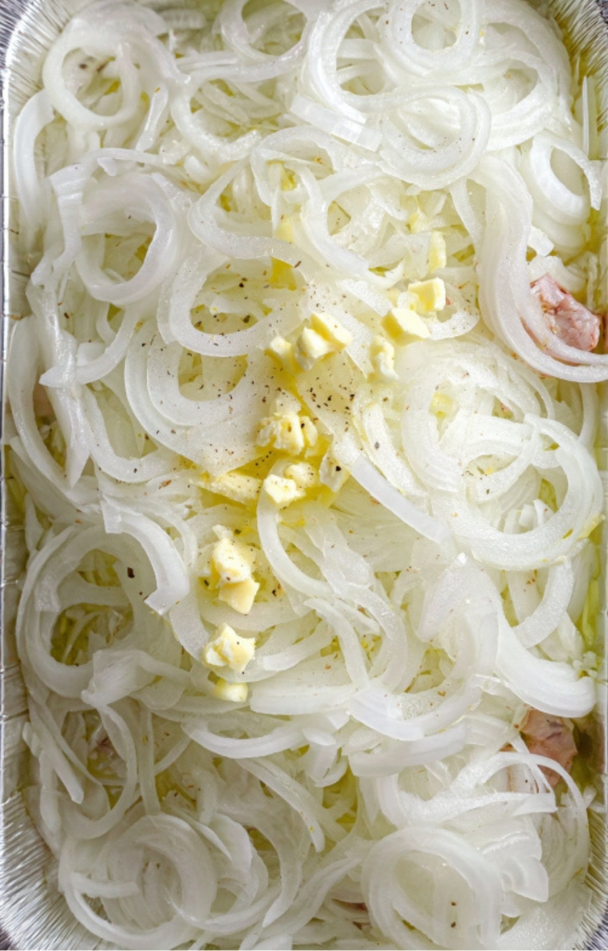 A foil tray filled with a thick layer of sliced white onions topped with pats of butter and black pepper.