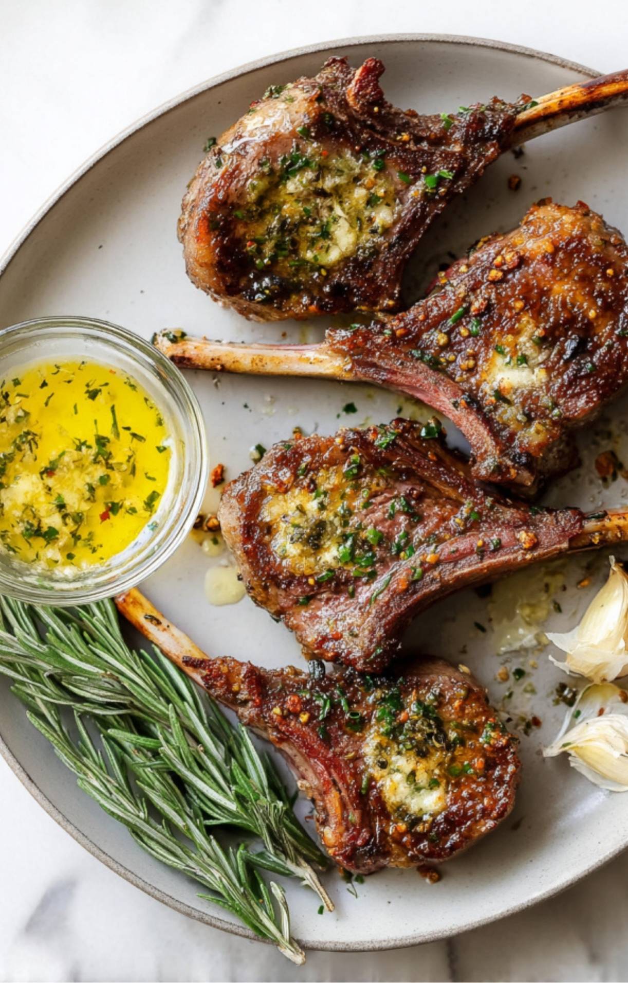 A top-down shot of four seared lamb chops on a grey plate, served with a small glass bowl of garlic herb butter, fresh rosemary sprigs, and whole garlic cloves on a marble surface.