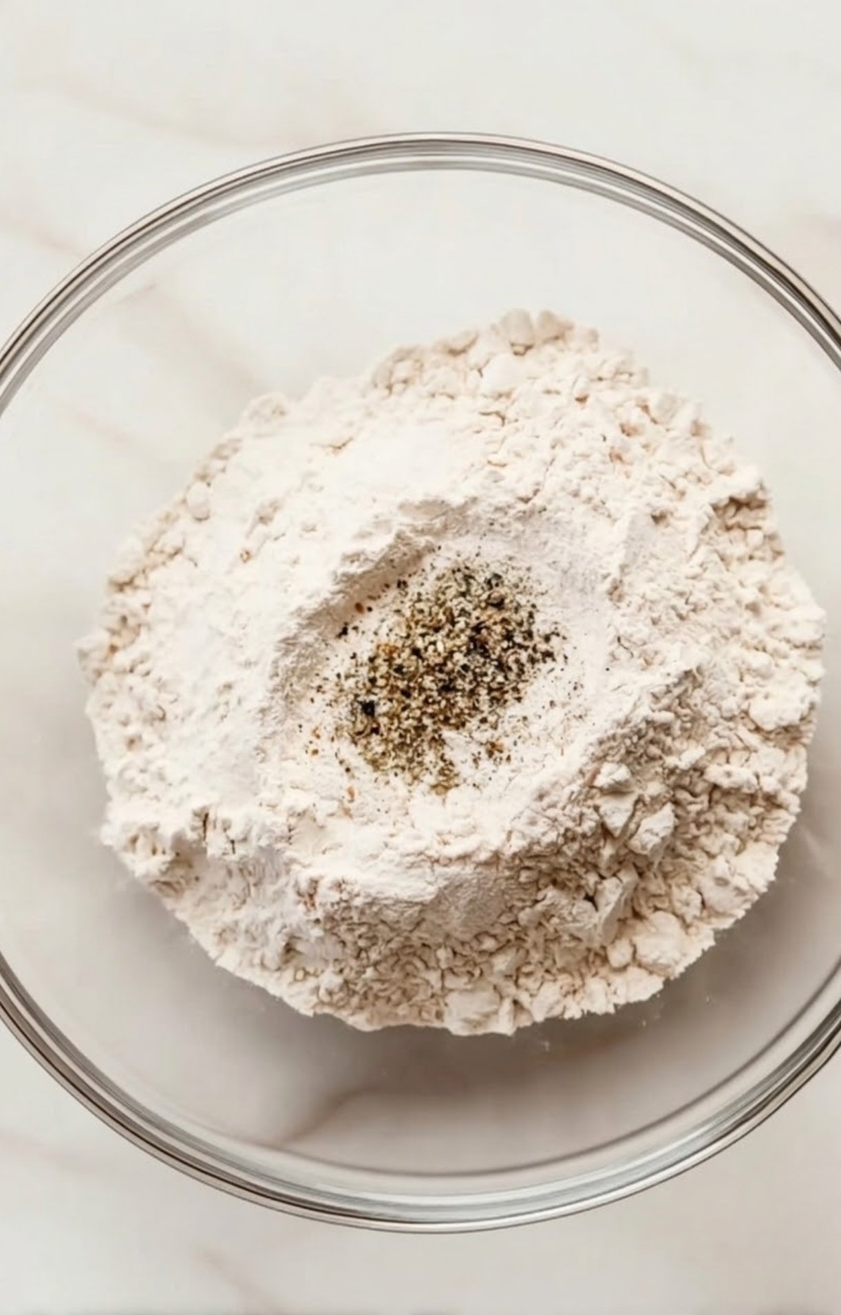 A glass bowl containing white flour topped with salt and cracked black pepper, ready for dredging Beef Cheeks.