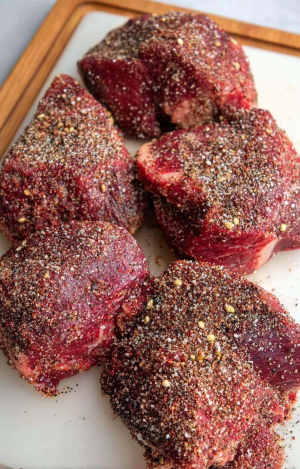 Several raw beef cheeks heavily coated in a coarse salt and black pepper dry rub resting on a white cutting board.