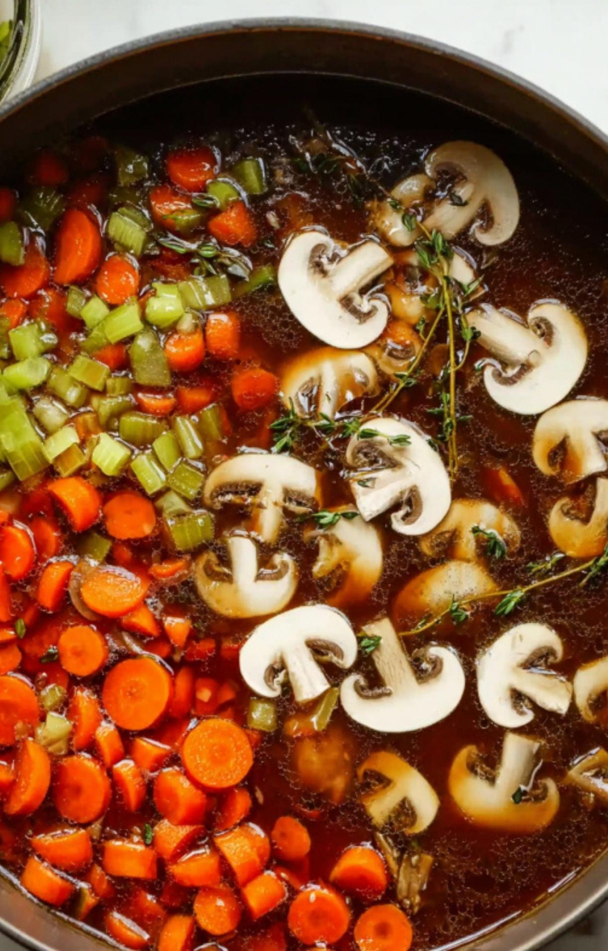 A large pot containing Beef Cheeks braising in a rich liquid with sliced mushrooms, carrots, celery, and fresh thyme sprigs.