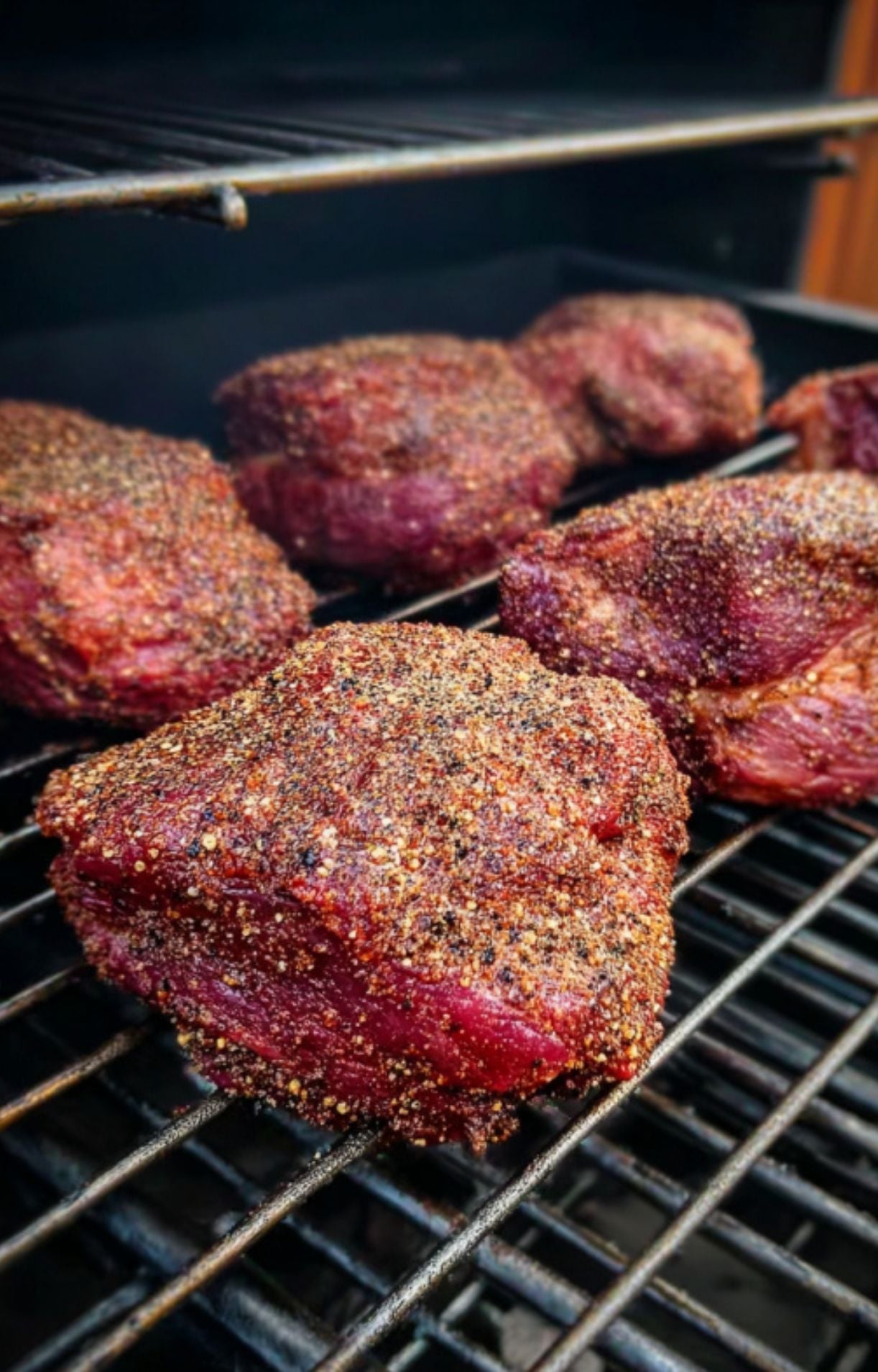 Raw, seasoned beef cheeks placed on metal grill grates inside a smoker, beginning the slow-cooking process for smoked beef cheeks.