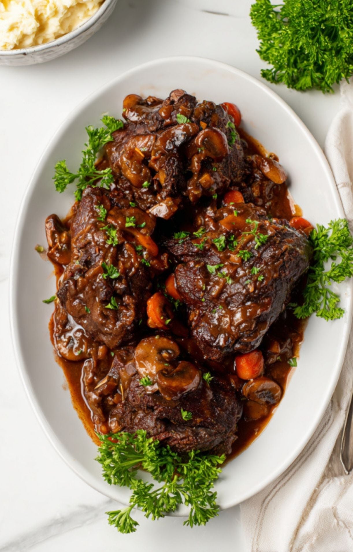 A white oval serving platter filled with tender, braised Beef Cheeks smothered in a thick mushroom gravy and garnished with fresh parsley.
