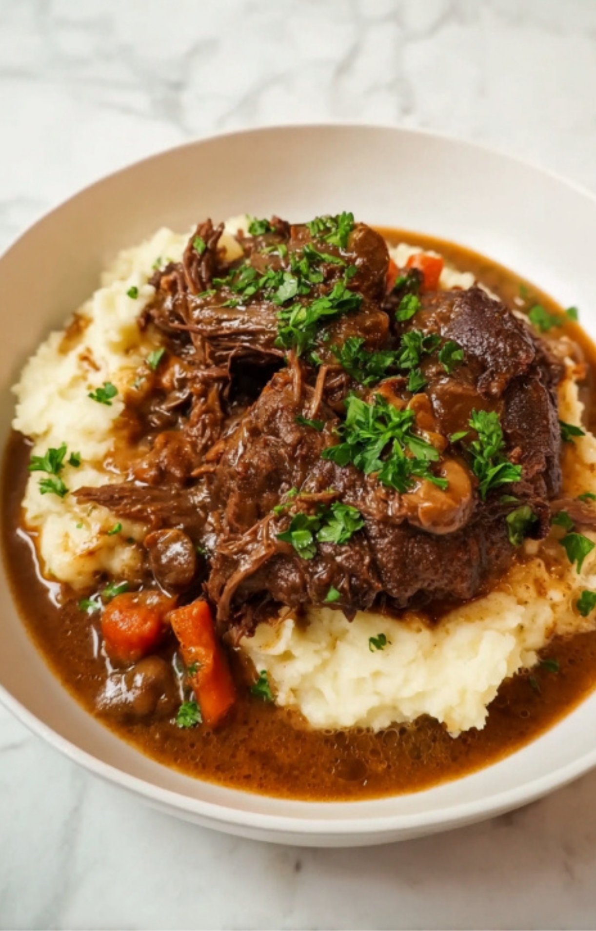 A white bowl featuring a tender, fall-apart braised Beef Cheek served over creamy mashed potatoes with a side of carrots.