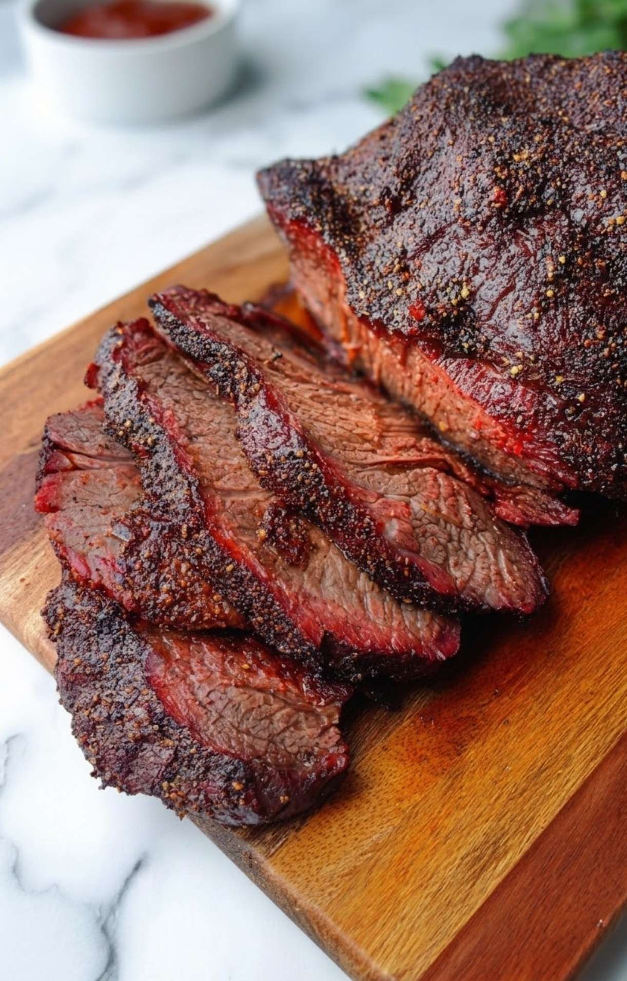 Slices of smoked beef cheeks resting on a wooden cutting board, showcasing a dark, seasoned bark and a prominent pink smoke ring with a small bowl of sauce in the background.