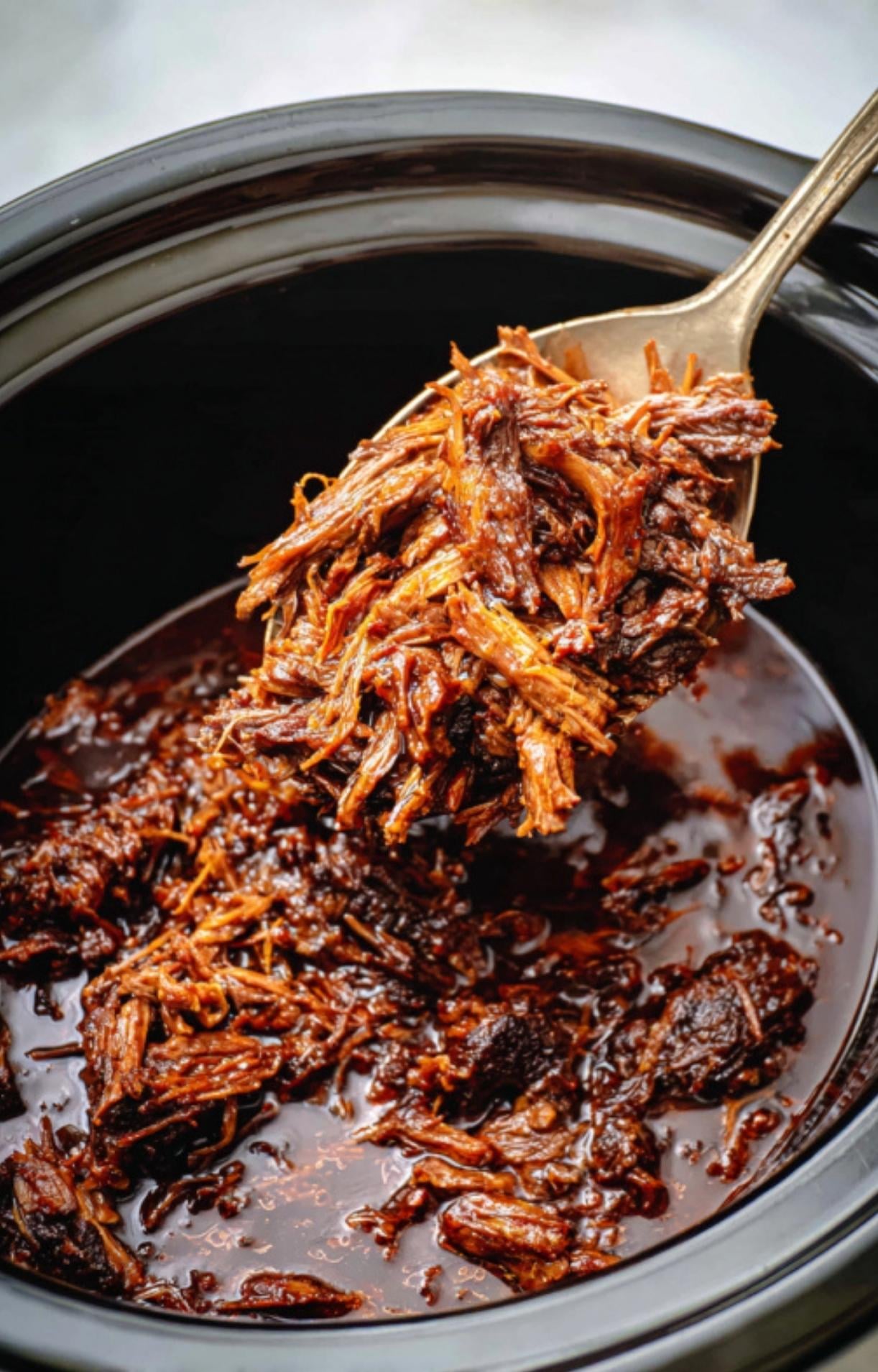 A gold spoon lifting a heap of succulent, fork-tender shredded Beef Cheek Barbacoa from a dark slow cooker pot.