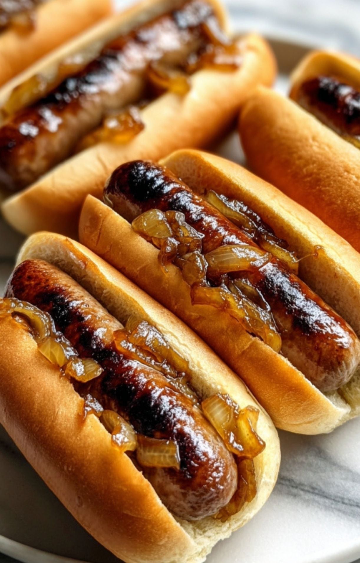 A row of four beer brats perfectly browned and nestled in buns, topped with golden-brown onions cooked in beer.