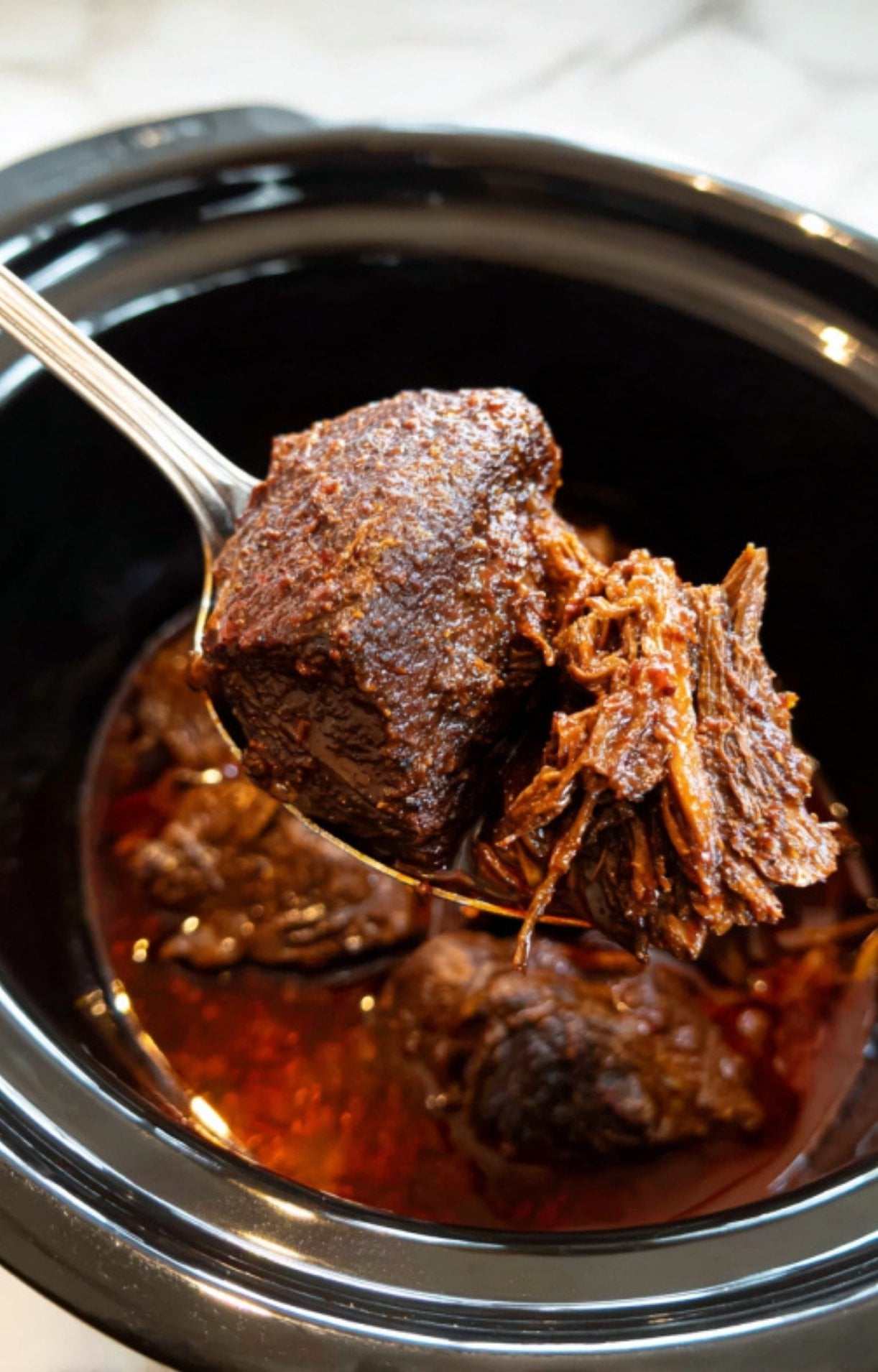 A silver spoon lifting a large, succulent chunk of braised beef cheek and some shredded meat from a dark slow cooker pot filled with rich red consommé.