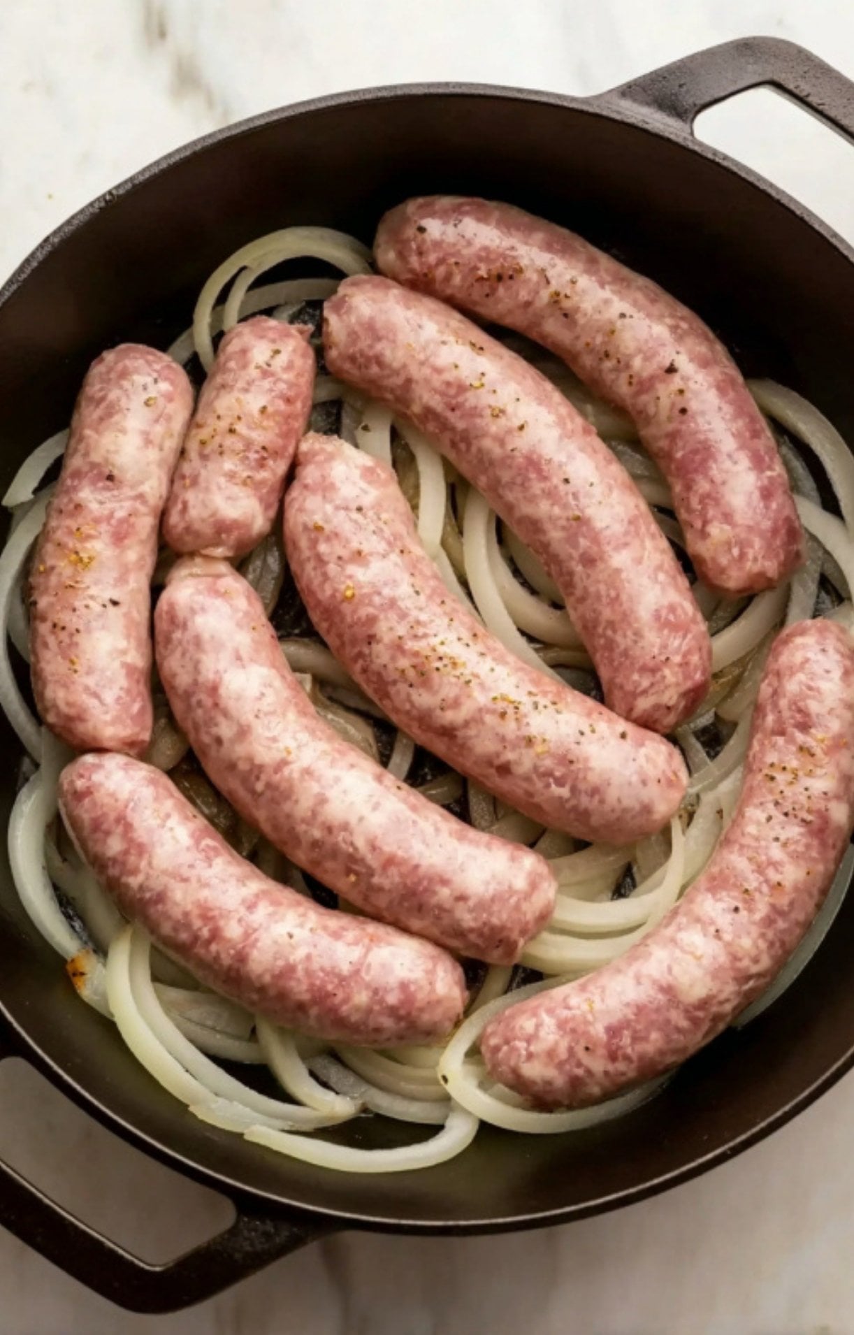 Raw bratwurst sausages seasoned with pepper and nestled on a bed of sliced white onions in a black cast iron skillet.