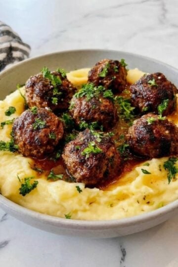 A bowl of golden mashed potatoes topped with six browned air fryer beef meatballs and fresh parsley.
