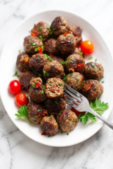 A white oval platter filled with browned lamb kofta meatballs garnished with fresh parsley and cherry tomatoes on a white marble surface.