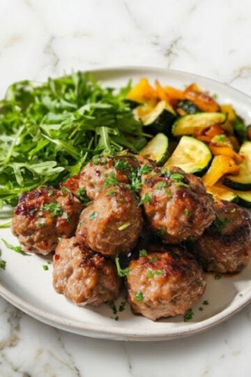A full serving of Air Fryer Pork Meatballs on a white ceramic dish with a side salad and roasted vegetables, styled on a gold-veined marble background.