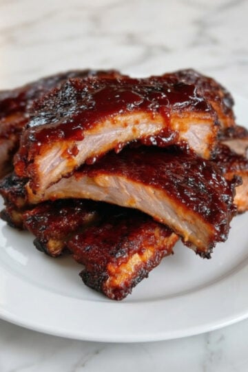 A stack of juicy, BBQ-glazed baby back ribs served on a white plate over a clean white marble background.