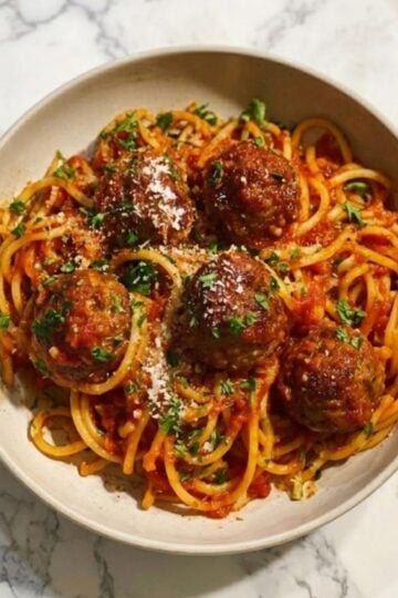 A white bowl filled with spaghetti and tomato sauce topped with five large air-fried meatballs, fresh parsley, and grated parmesan cheese.