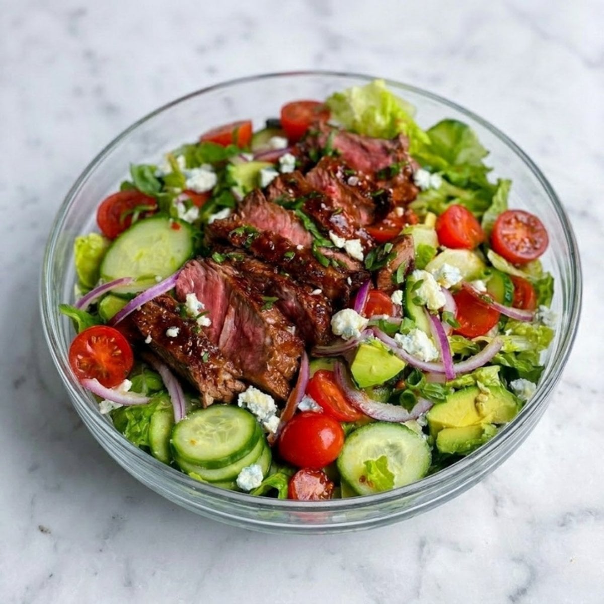 A vibrant glass bowl filled with a fresh Steak Salad Recipe featuring sliced medium-rare steak, cherry tomatoes, cucumbers, red onions, avocado, and blue cheese crumbles.