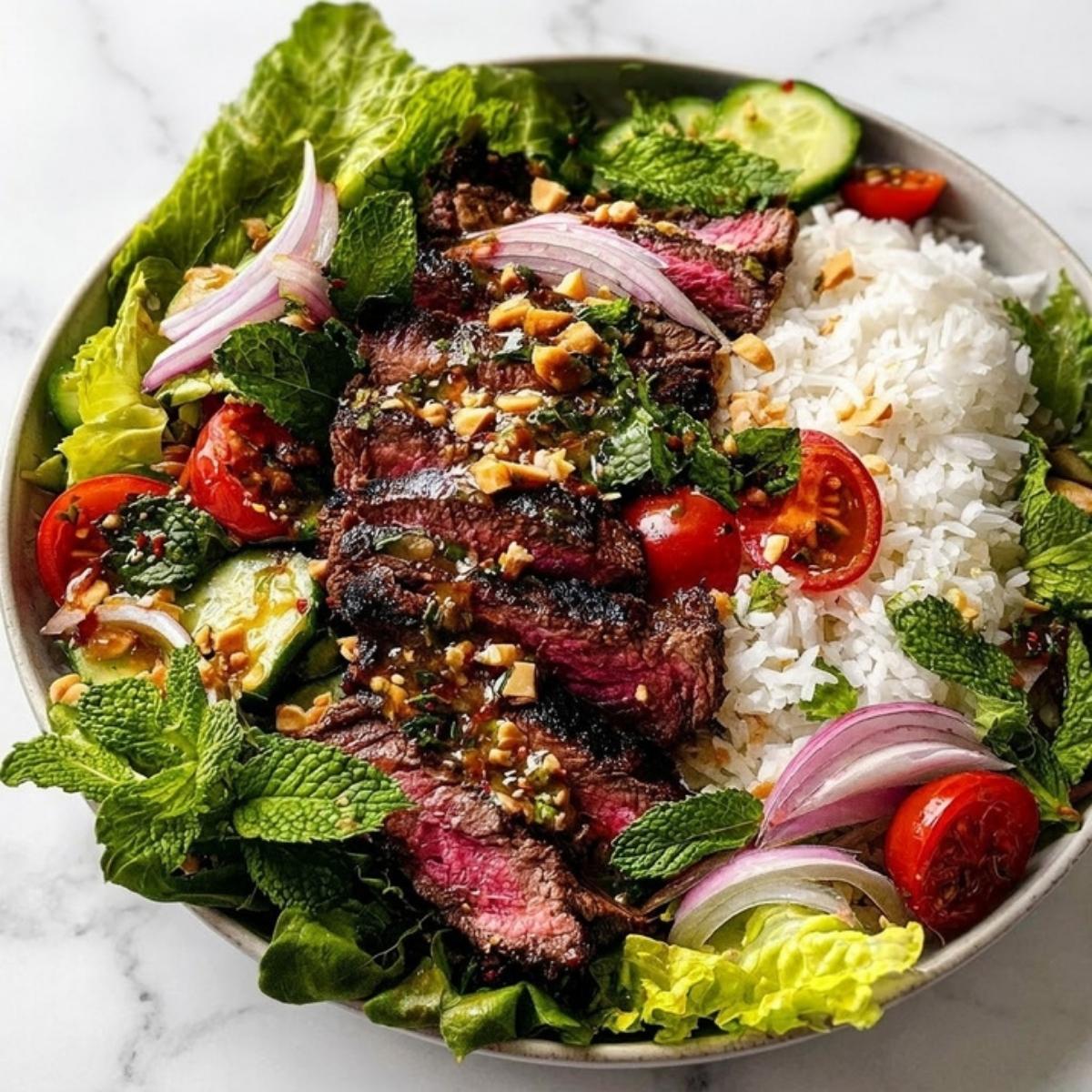 An overhead shot of a colorful Thai steak salad garnished with toasted peanuts, lime, and fresh herbs in a large serving bowl.