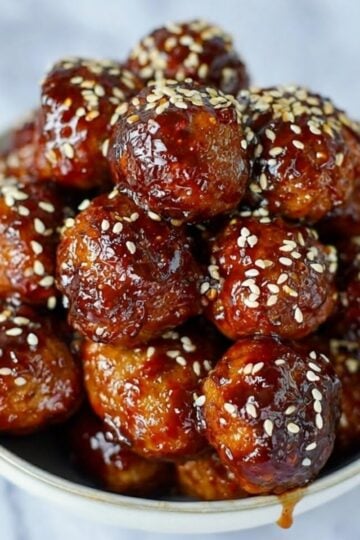 A large bowl of cooked frozen meatballs in air fryer coated in a dark, glossy glaze and topped with white sesame seeds.