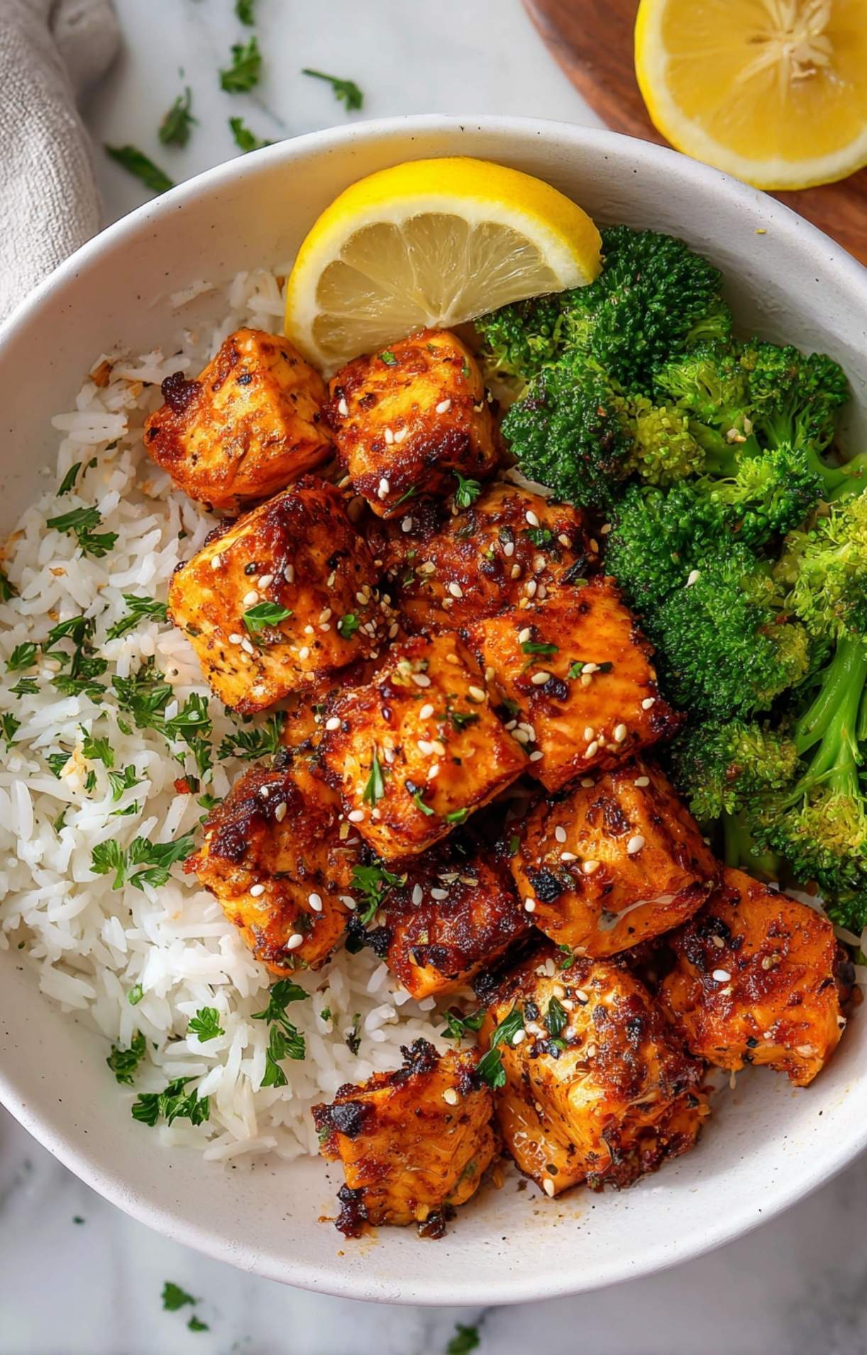 A nutritious meal bowl featuring golden-brown Air Fryer Salmon Bites served alongside fluffy white rice topped with parsley and steamed green broccoli, finished with a lemon squeeze.