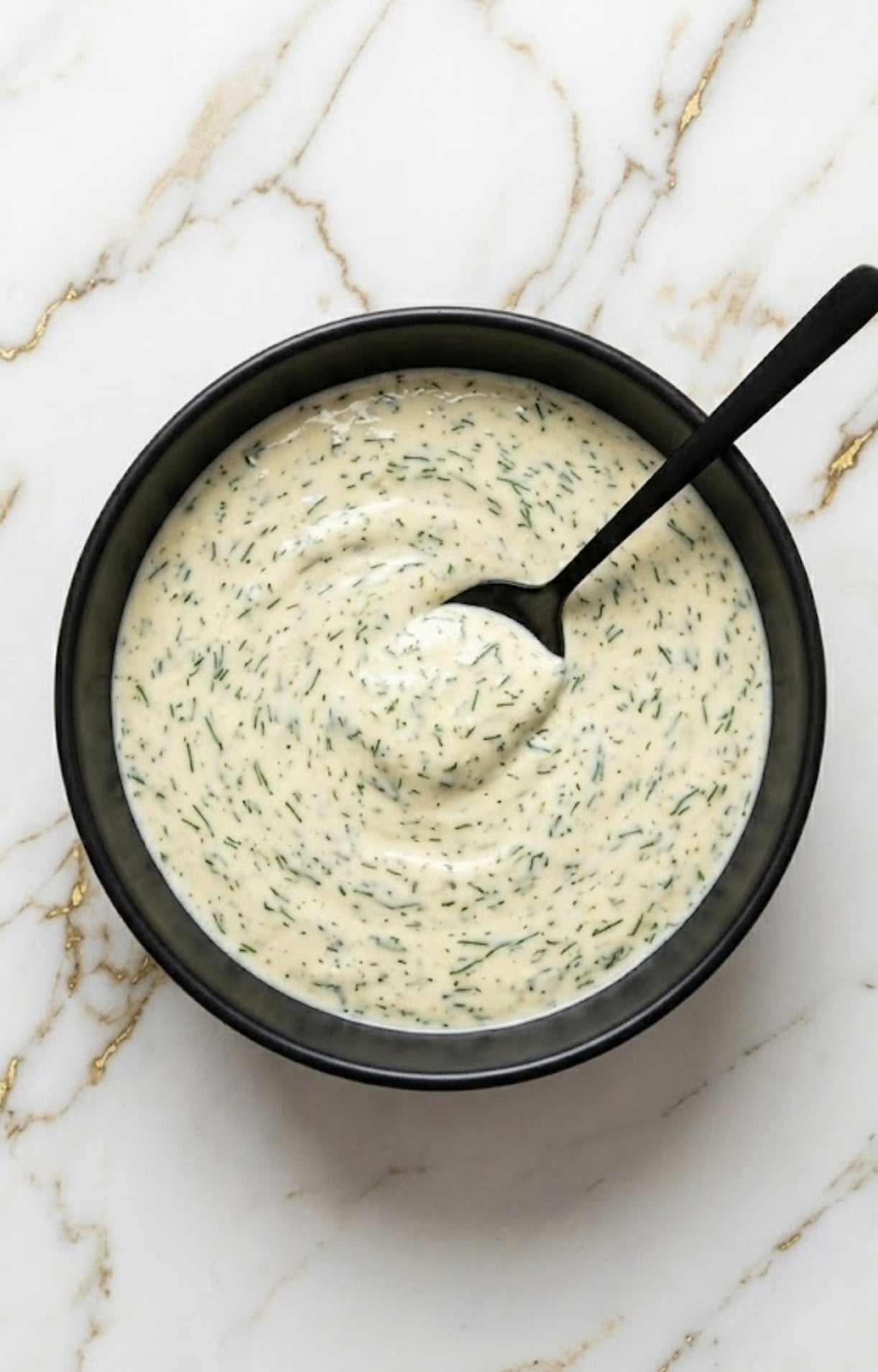 A black bowl filled with a creamy white herb sauce being stirred with a black spoon.