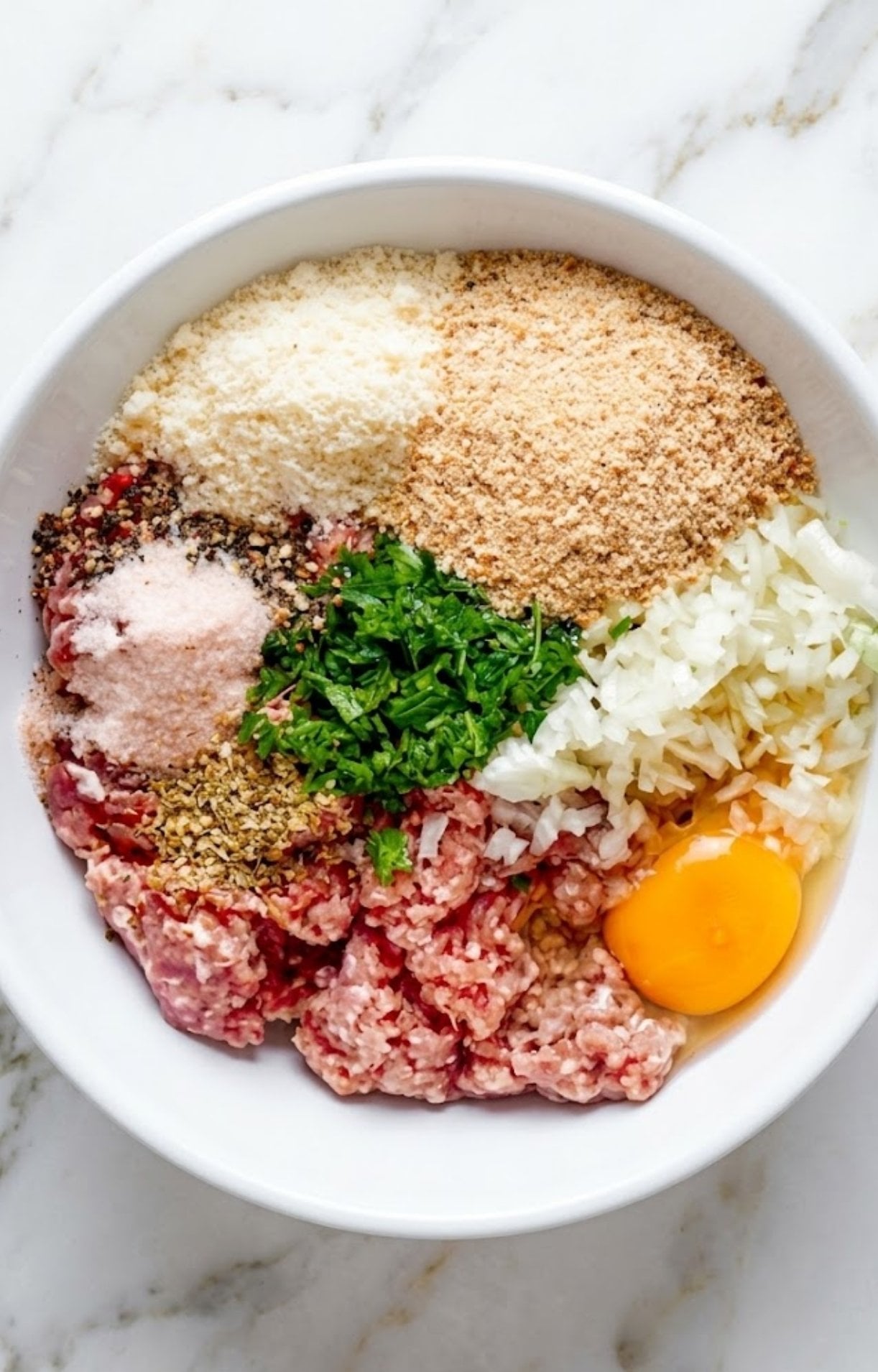 A white bowl containing ground beef, an egg, breadcrumbs, parmesan, diced onions, and herbs.