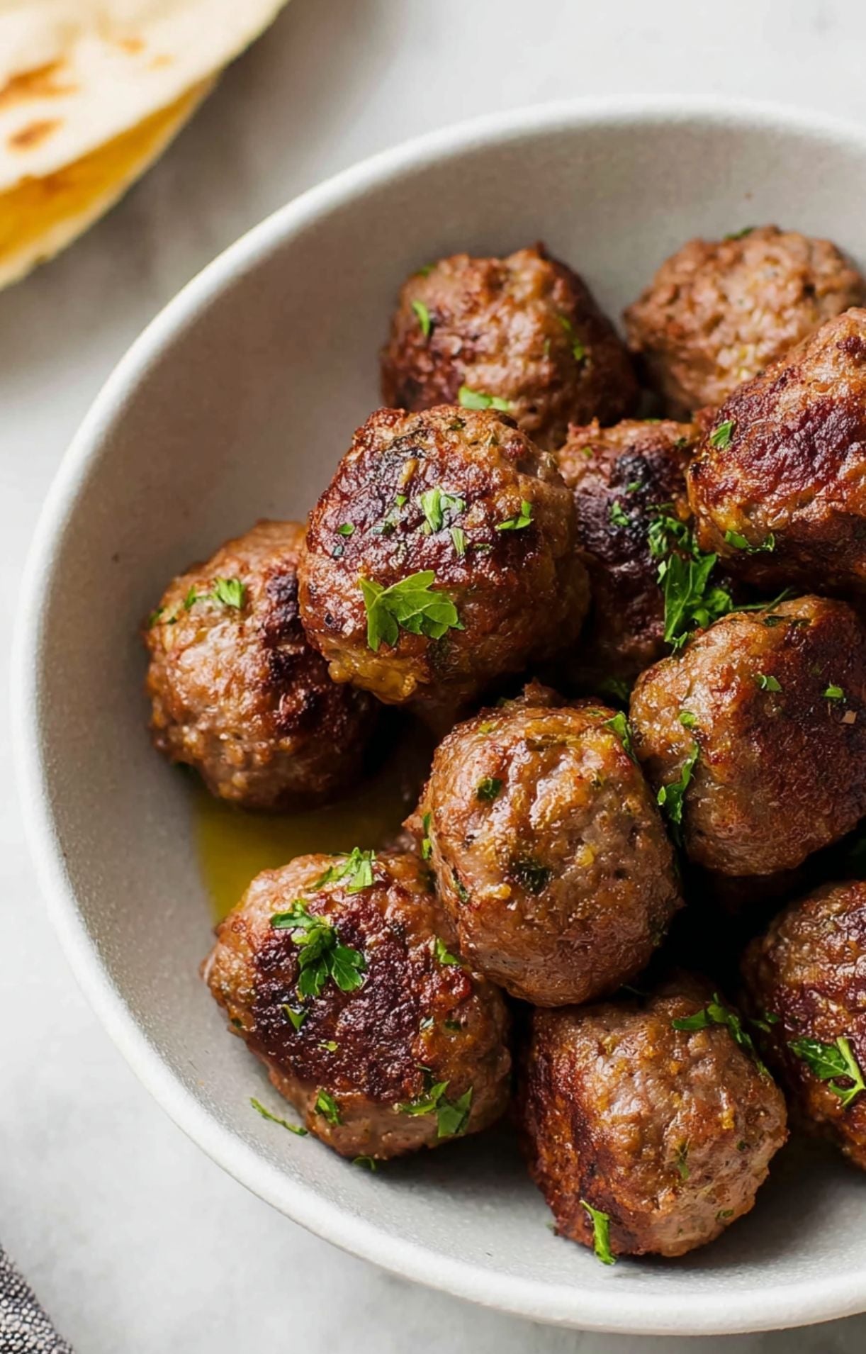 A close-up of cooked, crispy lamb meatballs in a grey ceramic bowl served alongside warm flatbread.