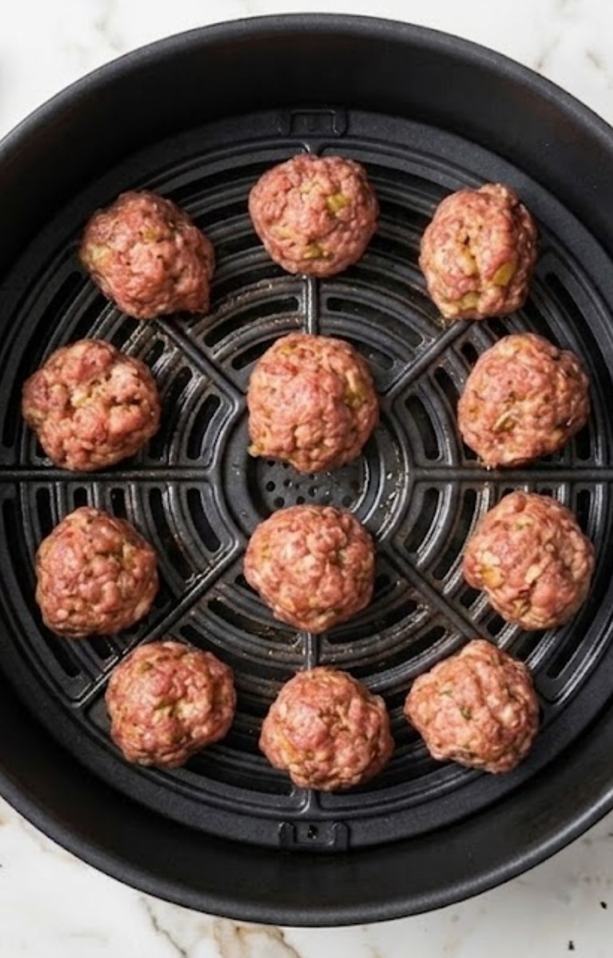 Raw pork meatballs arranged in a single layer inside a black air fryer basket sitting on a white marble countertop.