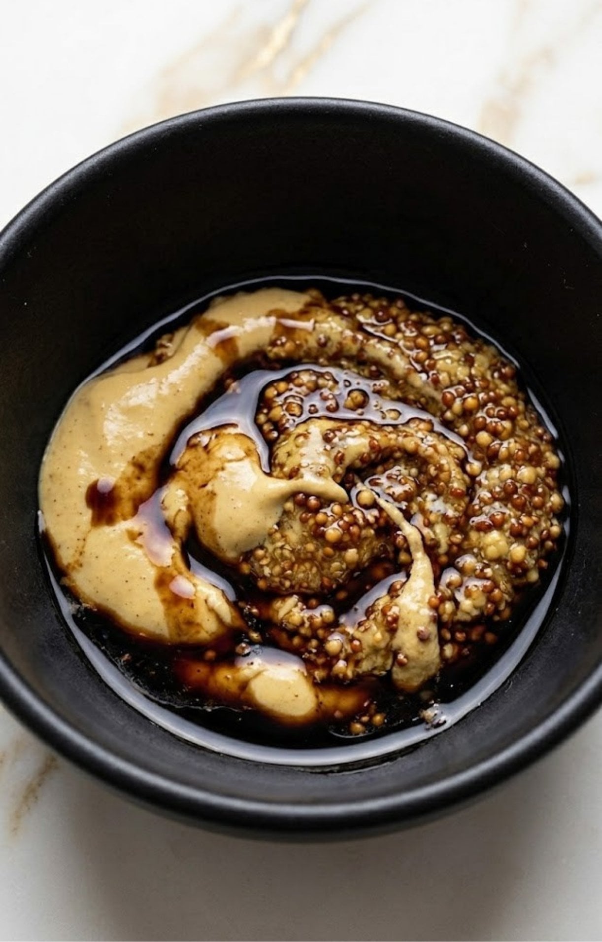 A black bowl containing a swirl of smooth Dijon mustard, grainy mustard, and dark balsamic vinegar.