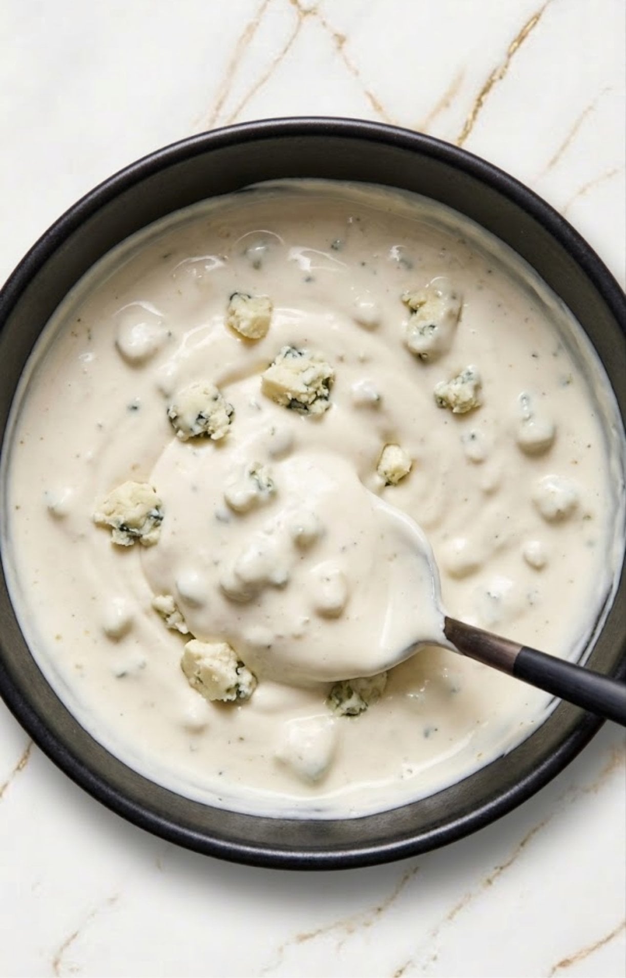 A close-up of a black bowl filled with thick, creamy white dressing topped with chunky blue cheese crumbles.