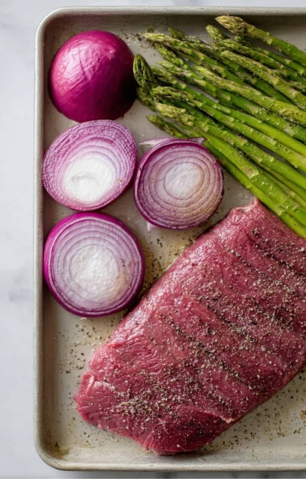 A baking sheet holding a raw flank steak seasoned with salt and pepper, fresh asparagus spears, and thick slices of red onion.