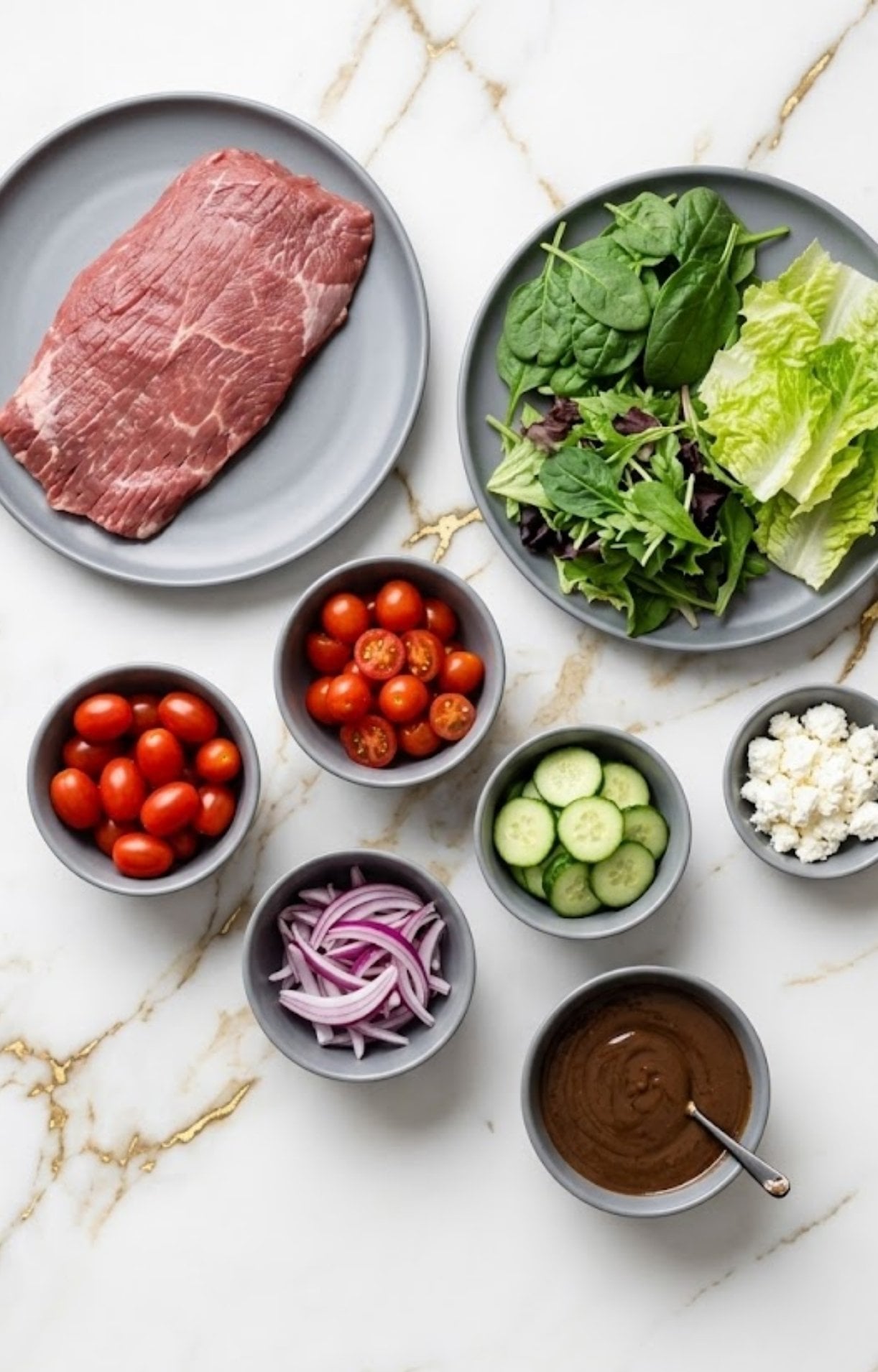 A flat lay of raw ingredients for a Healthy Steak Salad Recipe, including a raw flank steak, fresh greens, cherry tomatoes, sliced cucumbers, red onions, feta cheese, and a balsamic vinaigrette.